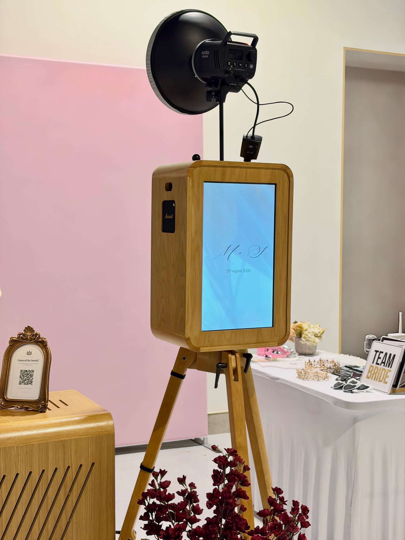 The Booth Royale oak photo booth on a wooden tripod at a wedding, with studio flash, blush backdrop, custom welcome screen and matching print station.